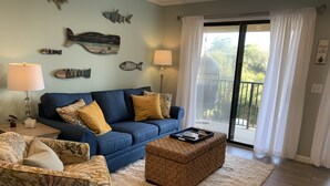 Un salón con sofá azul, sillón estampado, reposapiés de mimbre y un amplio ventanal con vistas a zonas verdes.