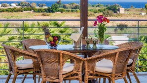 Une table en osier avec des chaises et une table avec un plateau en verre, un saladier rempli de fruits et un vase de fleurs sur un balcon donnant sur la mer.