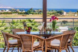 Mesa de vime com cadeiras e uma mesa com tampo de vidro, uma cesta de frutas e um vaso com flores numa varanda com vista para o mar.