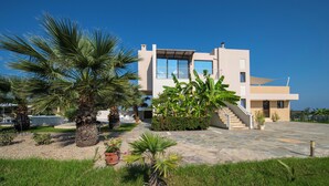Une maison moderne à deux étages, dotée d’une terrasse en pierre, de palmiers et surplombant un ciel d’un bleu éclatant.