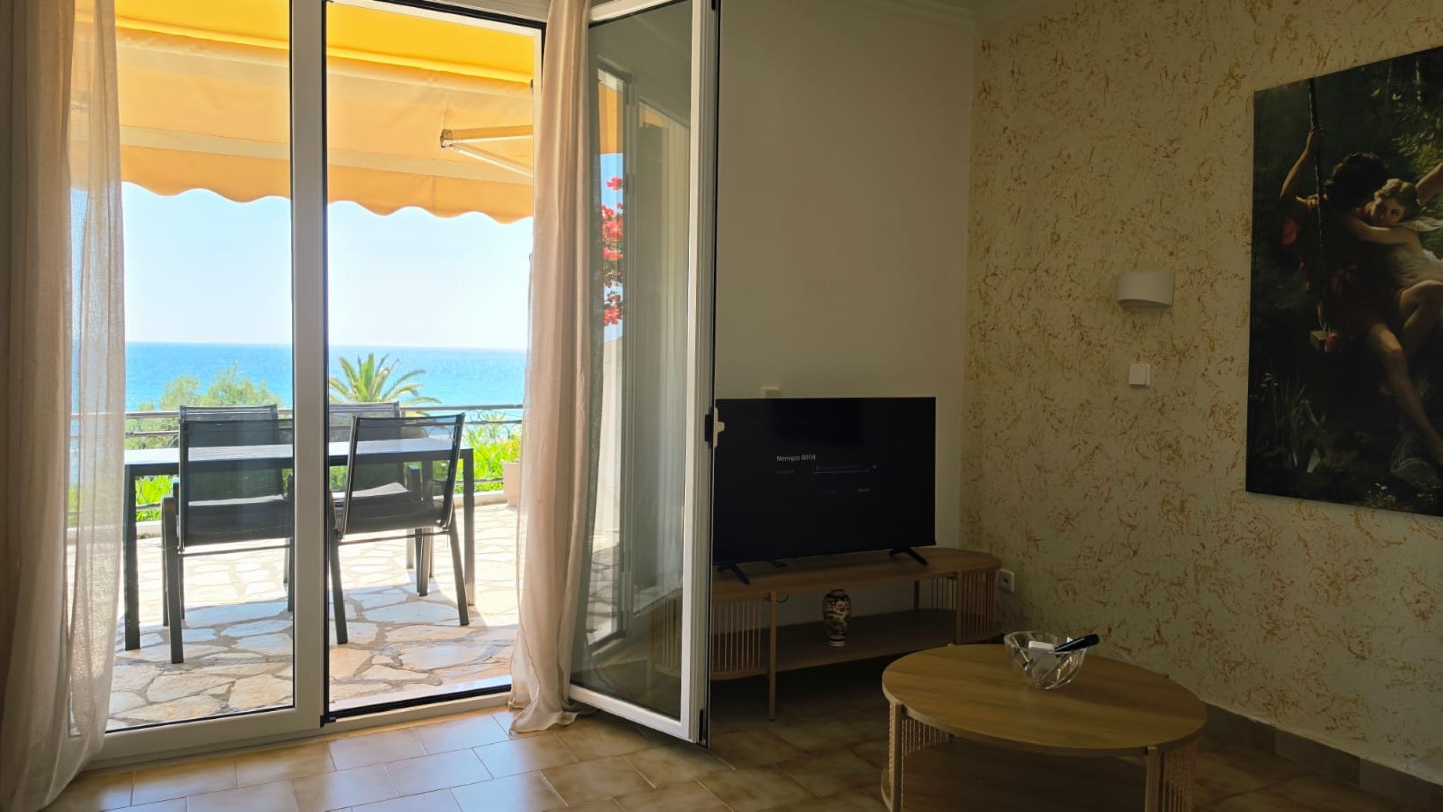 Una habitación con puerta de vidrio corrediza a un balcón, televisión, una mesita y un cuadro en la pared.