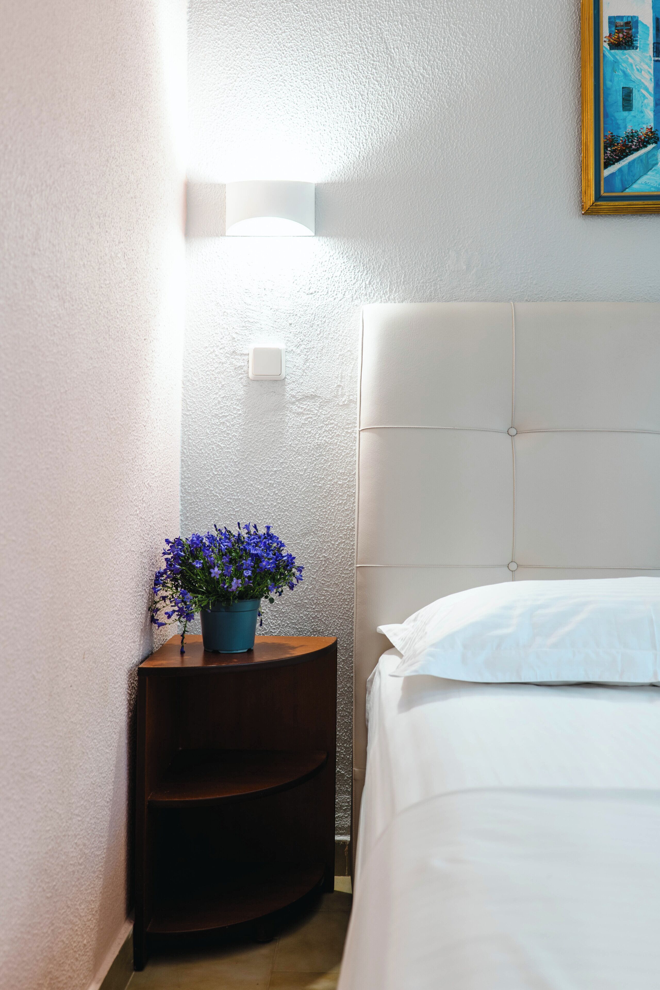 A hotel room with a bed, a nightstand with a blue flower pot, and a painting on the wall.