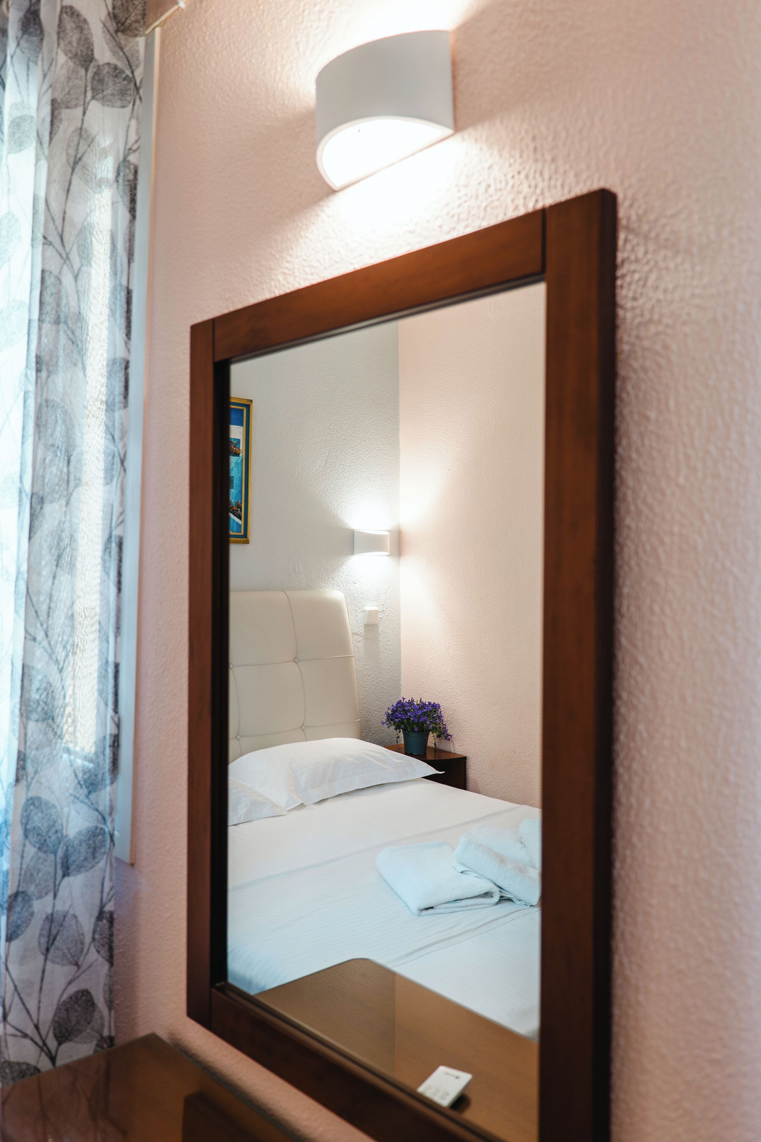 A hotel room with a large mirror, a bed with white linens, a nightstand with a lamp, and a window with curtains.