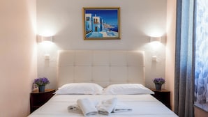 Una camera d'albergo con un letto, cuscini, un quadro, comodini con fiori e una tenda.
