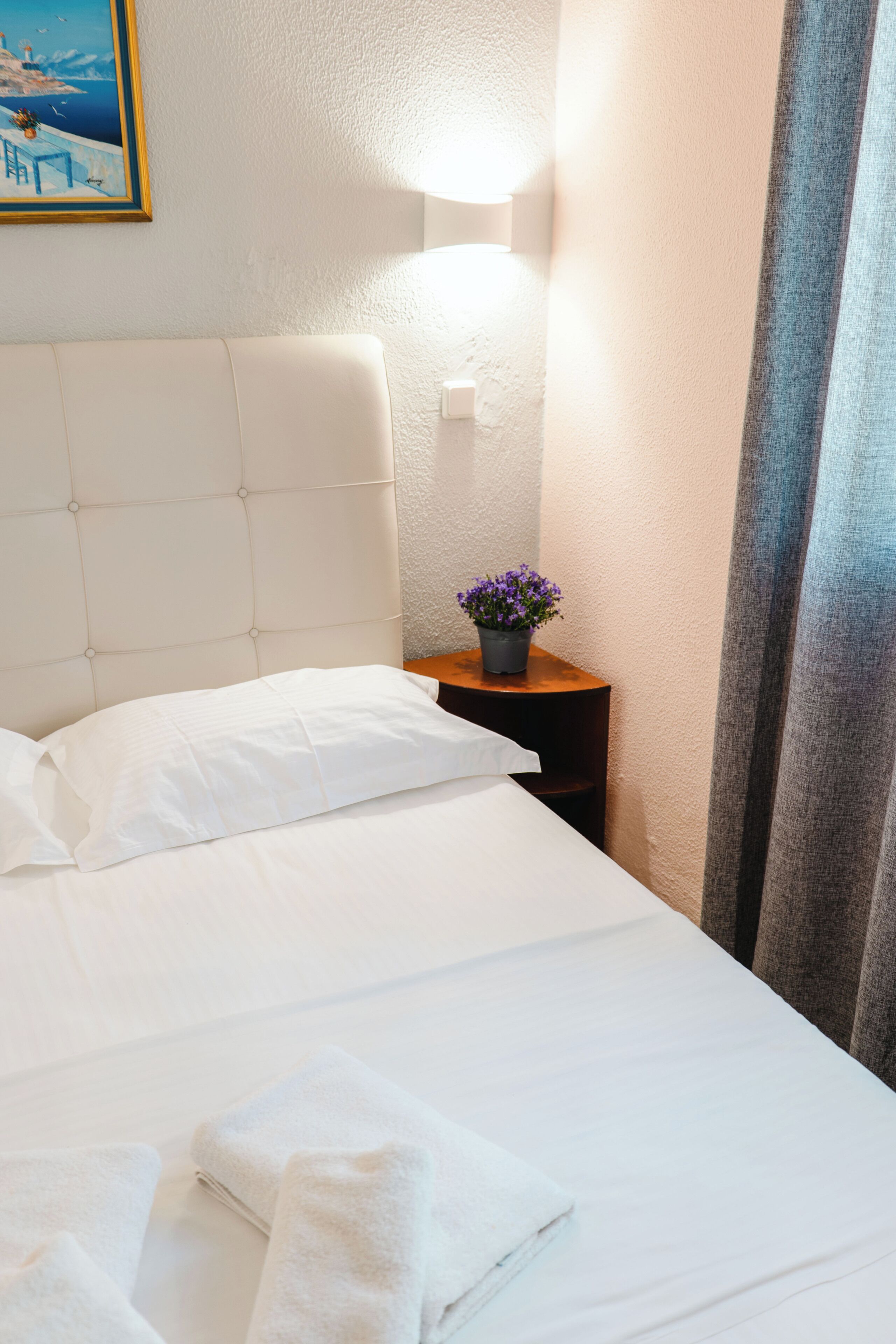 A neatly made bed with white linens, a small bedside table with a potted plant, and a wall-mounted light fixture.