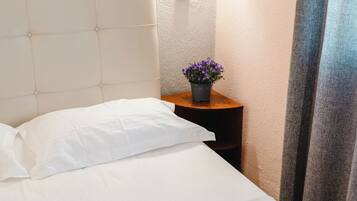 A neatly made bed with white linens, a small bedside table with a potted plant, and a wall-mounted light fixture.