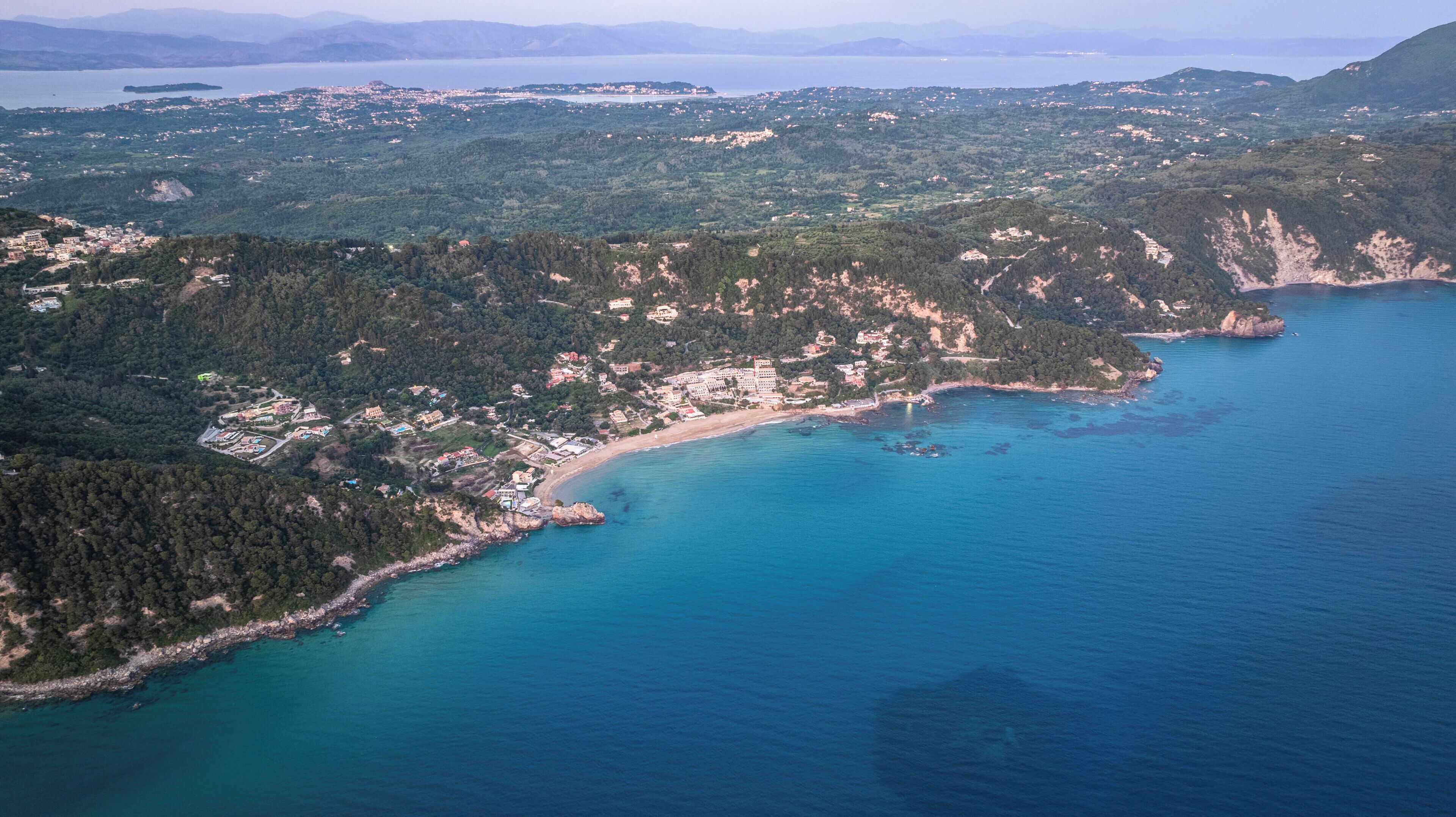 Vista aérea de uma área costeira com praia, edifícios e uma encosta arborizada.