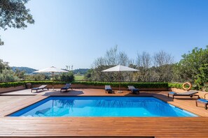 A swimming pool with lounge chairs and umbrellas, surrounded by greenery and hills.