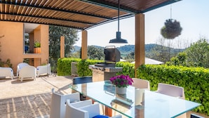 A glass-top table with chairs, a grill, and a hanging planter in an outdoor patio area with a view of the hills. -  Charming Country House with Large Garden with Mountain View, Pool, Wi-Fi and Terraces (Artá)