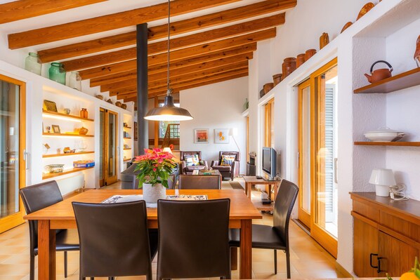 A dining area with a wooden table, chairs, a hanging light fixture, and shelves with kitchenware.