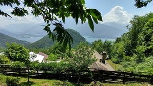 A rural house with a sloped roof, surrounded by lush greenery and a view of a lake and mountains. - Cosy apartment for 3 guests with WIFI, TV, patio and pets allowed (Peglio)