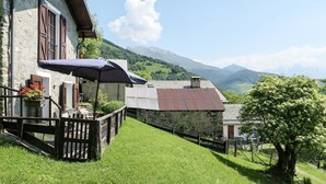 A rustic stone building with a wooden deck and a large umbrella, set against a backdrop of green hills and mountains.