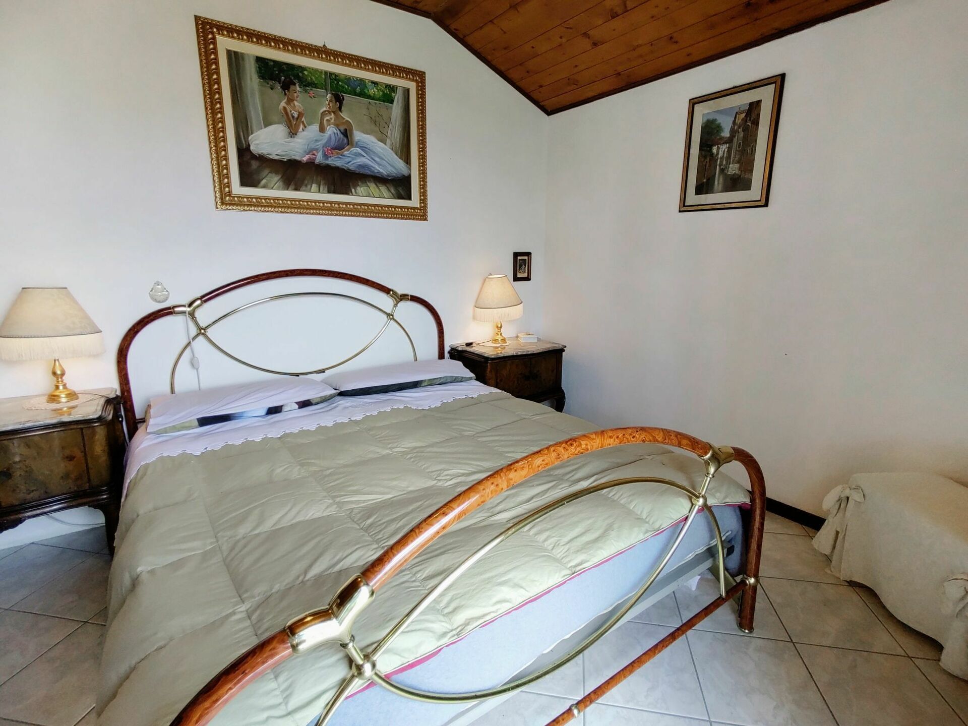A bedroom with a wooden bed frame, a white bedspread, two bedside tables with lamps, and two framed pictures on the walls.