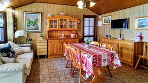Ein gemütliches Zimmer mit Holzbalkendecke, einem Esstisch mit rot-weiß kariertem Tischläufer, einem Holzschrank und einem Fernseher an der Wand.