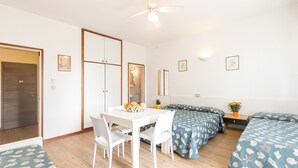 A room with a white table and chairs, two beds with matching bedding, a ceiling fan, and a small table with a vase of flowers. - Yachting Residence (Lignano Sabbiadoro)