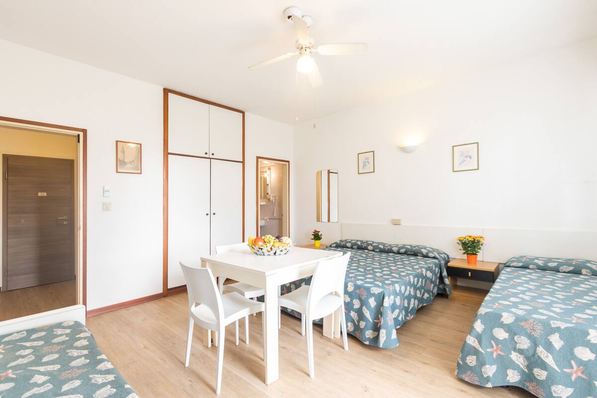 Een kamer met een witte tafel en stoelen, twee bedden met passend beddengoed, een plafondventilator en een kleine tafel met een bloevas.
