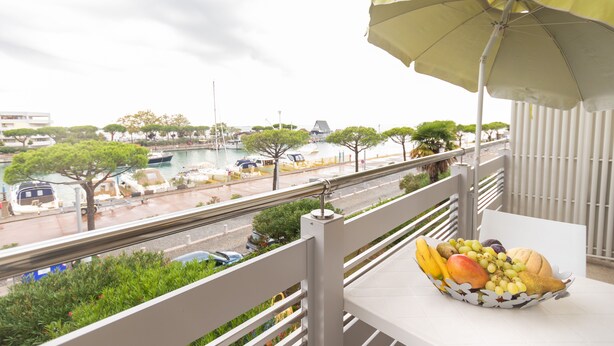 A balcony with a table holding a fruit basket, overlooking a marina with boats and trees. - Yachting Residence (Lignano Sabbiadoro)