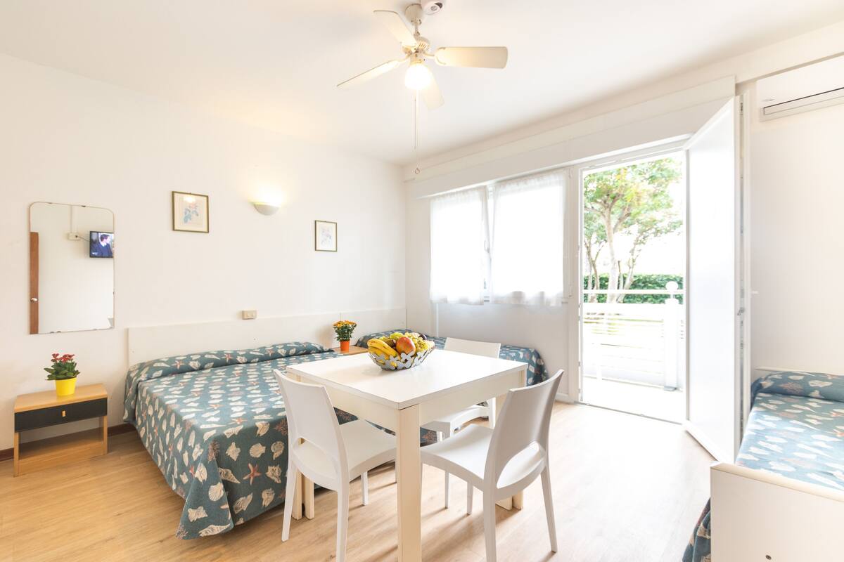 Een lichte kamer met een witte plafondventilator, een eettafel met stoelen, een bank en een raam met uitzicht op groen.