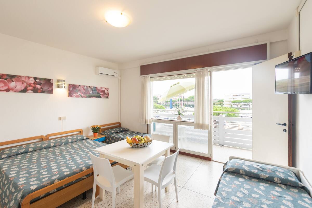 Een compacte kamer met een bed, een klein tafeltje met stoelen, een schaal fruit en een televisie.
