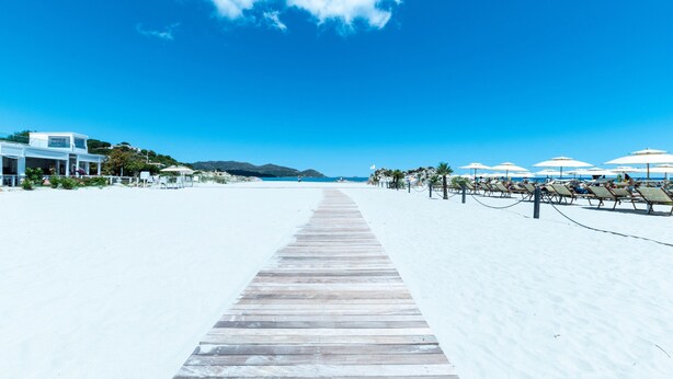 Une plage avec une promenade en bois menant à la mer, des parasols de plage et un bâtiment doté d’un balcon.