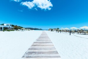 Una playa con un paseo de madera que conduce al mar, sombrillas de playa y un edificio con balcón.