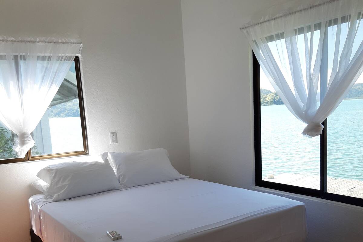 A bedroom with a bed, a window with sheer curtains, and a view of the water.