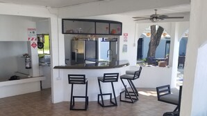 A kitchen area with a refrigerator, sink, and bar stools. There is a ceiling fan and a fire extinguisher on the wall. - Vista Castillo Hotel (Golfito)