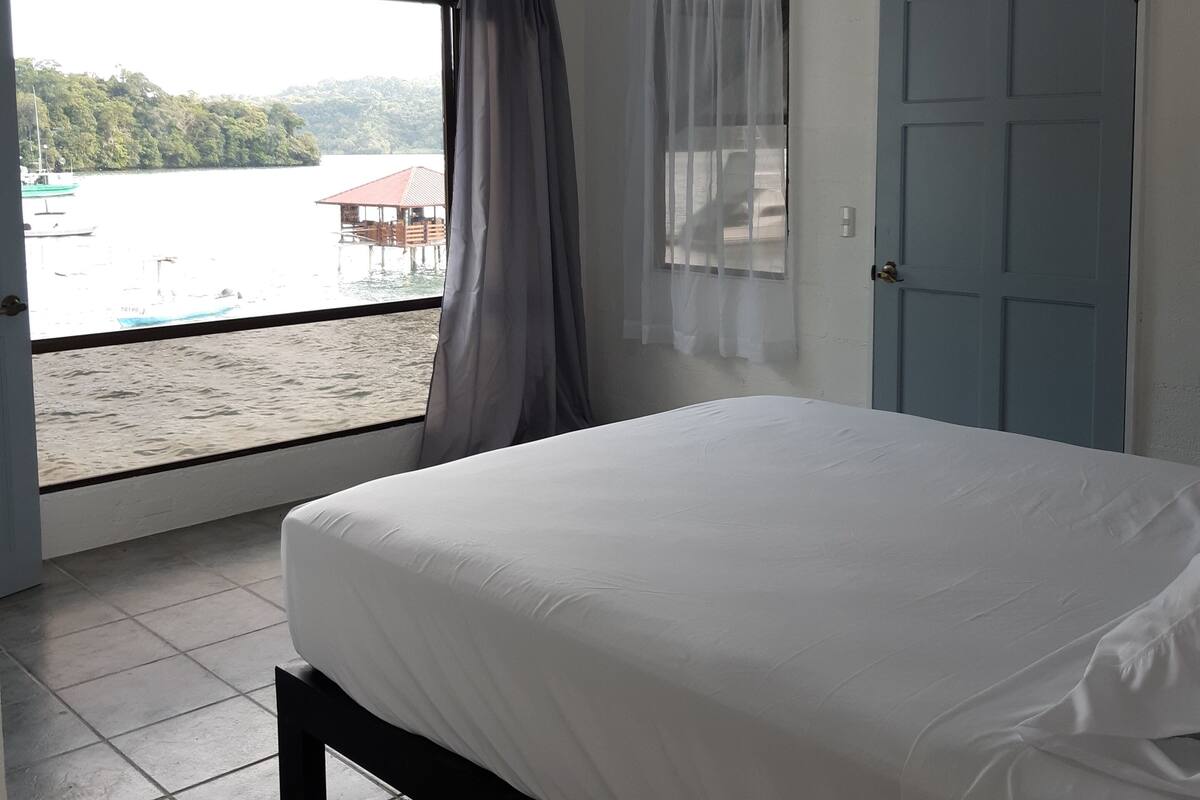 A bedroom with a bed, a window with curtains, a door, and an air conditioning unit.