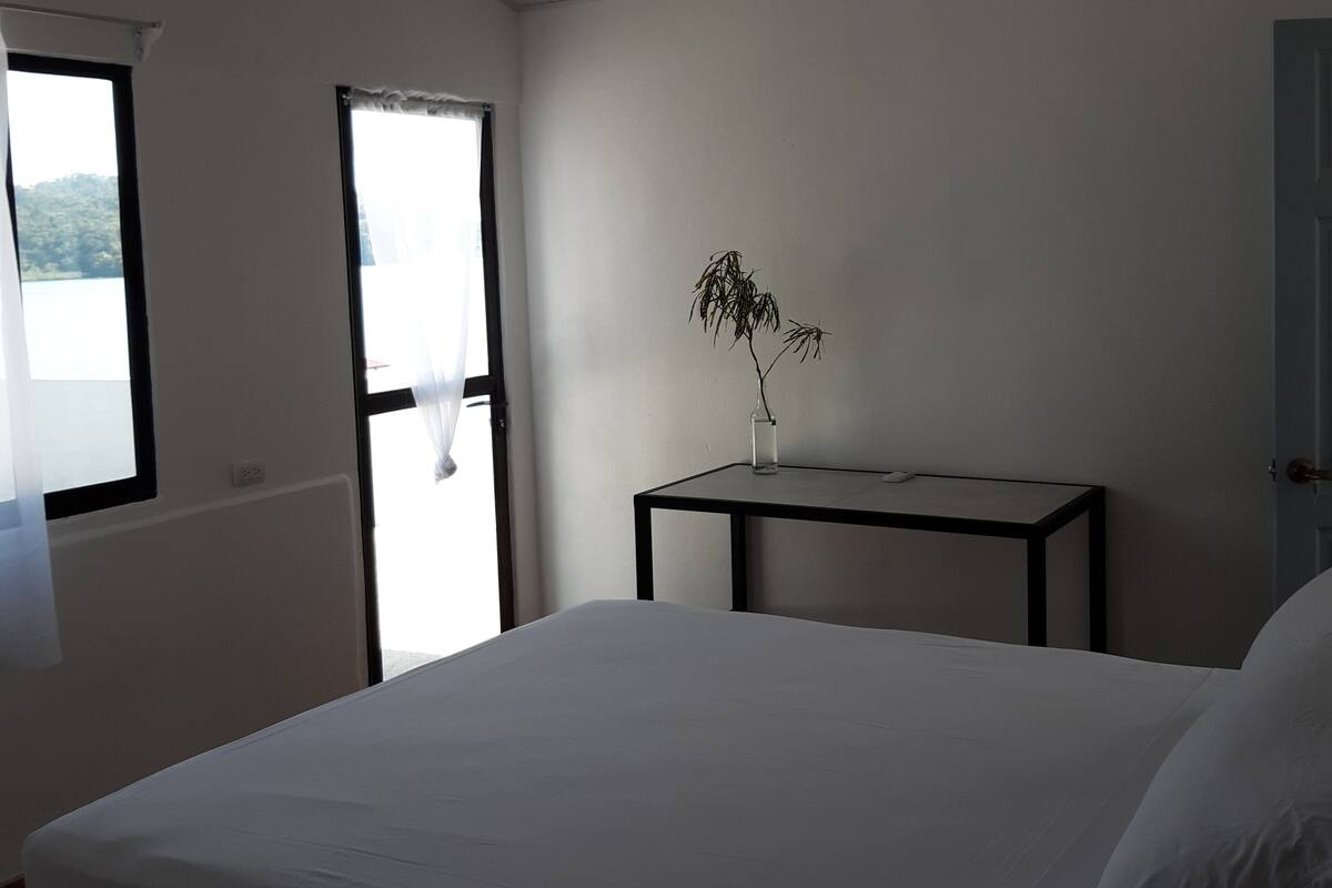 A bedroom with a large bed, a small table with a vase, and a hanging light fixture.