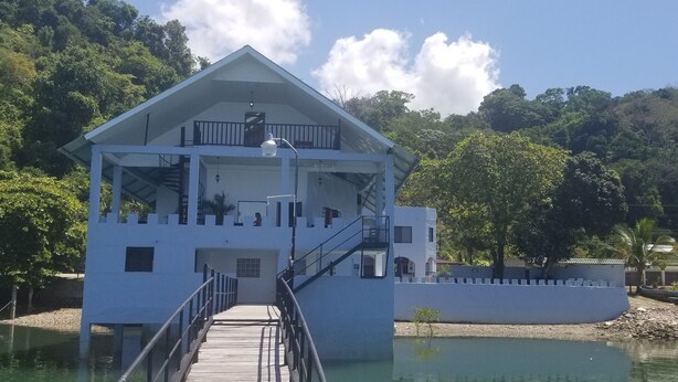 A white building with a balcony and a wooden walkway leading to it, surrounded by water and greenery. - Vista Castillo Hotel (Golfito)