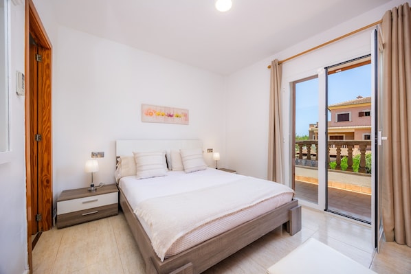 A bedroom with a bed, nightstands, a wall clock, a window with curtains, and a view of buildings outside.