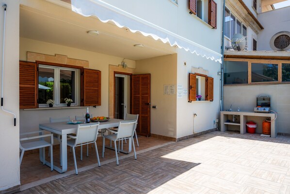 A patio with a table and chairs, a small kitchen area with a sink and a red bucket, and a window with shutters.