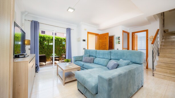 Una sala de estar moderna con un sofá azul, una mesa de centro de madera y un televisor sobre un mueble de madera. Se ve una escalera a la derecha y una puerta que da a otra habitación.