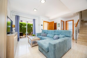 A modern living room with a blue sofa, a wooden coffee table, and a TV on a wooden stand. There is a staircase to the right and a door leading to another room.