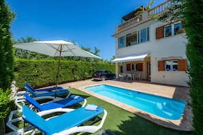 A pool area with lounge chairs and a white umbrella, a two-story house with a balcony, and a car parked nearby.