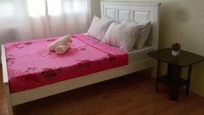 Een bed met een roze bloemenbedekking, witte kussens en opgerolde handdoeken. Er staat een klein tafeltje met een plantje naast het bed.