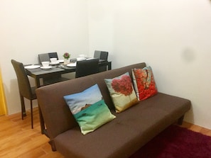 A living area with a brown sofa, two pillows, a dining table with chairs, and a small plant.