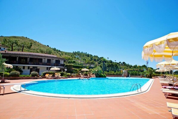 A swimming pool with lounge chairs and umbrellas, surrounded by a building with a tiled roof and greenery in the background. - Casa Vacanza Kair (Sperlonga)
