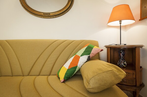 A mustard-colored sofa with a colorful geometric pillow, a wooden side table with a vase, and a wall-mounted mirror.