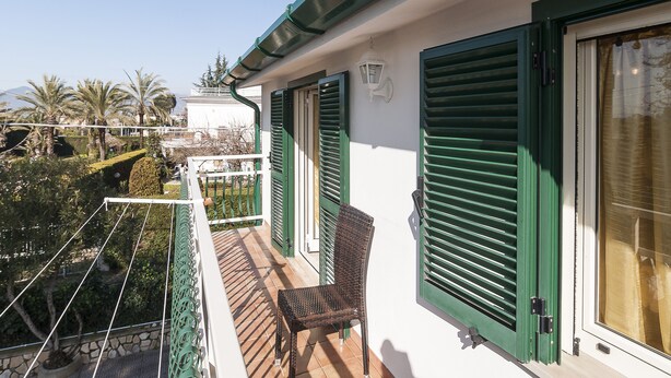 A balcony with a wicker chair, green shutters, and a view of a garden and mountains. - Casa Vacanza Argo (Sperlonga)