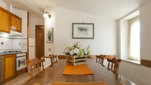 A dining room with a wooden table, chairs, a flower arrangement, and a kitchen in the background. - Casa Vacanza Argo (Sperlonga)