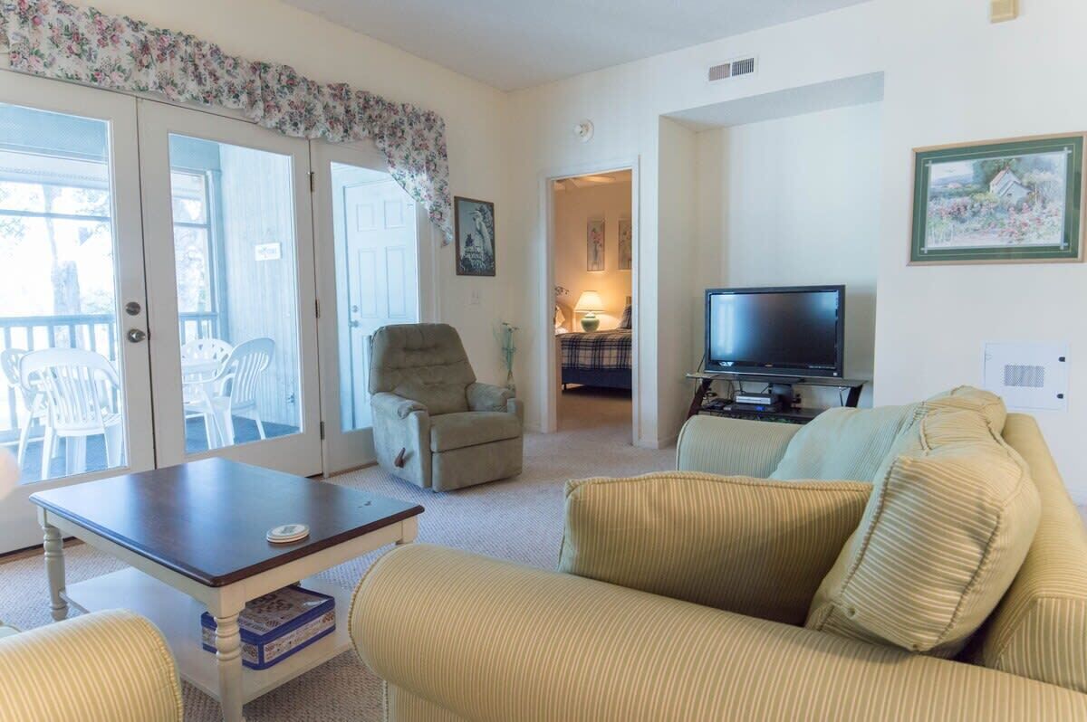 A living room with a sofa, armchair, and a television. There is a table and a chair in front of the sofa.