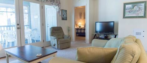 A living room with a sofa, armchair, and a television. There is a table and a chair in front of the sofa.