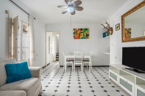 A living room with a ceiling fan, a TV, a dining table with chairs, a sofa with a cushion, and a patterned floor.