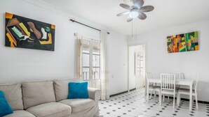 A room with a beige sofa, white chairs, a ceiling fan, and two abstract paintings on the walls.