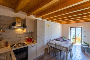 A modern kitchen with a gas stove, oven, and wooden ceiling beams. There is a dining area with a table and chairs, and a balcony with a view.