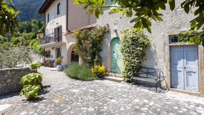 Une cour pavée de pierres, avec un banc, des plantes en pot et un bâtiment doté d’une porte verte et d’un balcon.