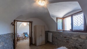 Une chambre avec un plafond en pente, une armoire en bois, un lit recouvert d’une couvre-lit à motifs, une chaise et une vue par une fenêtre ornée de rideaux en dentelle.