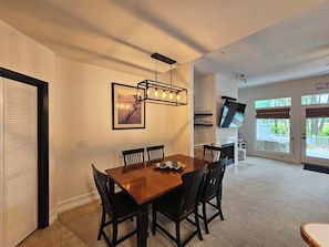 Un coin repas avec une table et des chaises en bois, un luminaire suspendu, un téléviseur fixé au mur et une vue sur l’extérieur par une porte.