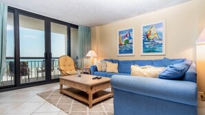 A living room with a blue sofa, a wooden coffee table, and a chair. There are two paintings on the wall and a balcony with a view of the sea. - Shoreham Towers I - C3 (North Myrtle Beach)